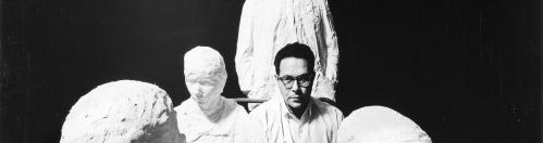 George Segal sits amongst plaster casts of bus riders. George is staring directly at the camera which is eye-level with him.